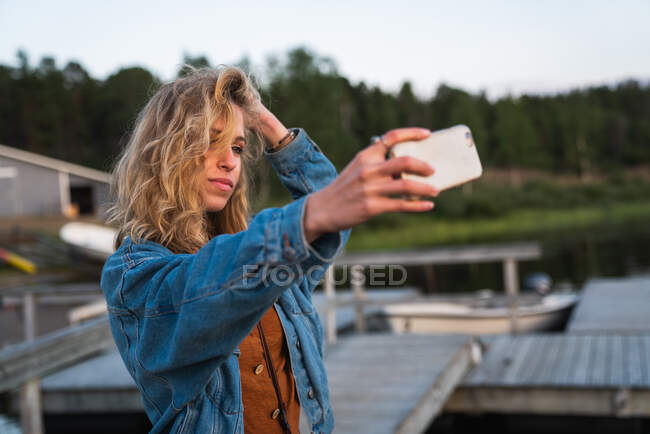Mujer atractiva joven con pelo claro en chaqueta de mezclilla de pie en la orilla del río y haciendo selfie con teléfono móvil - foto de stock