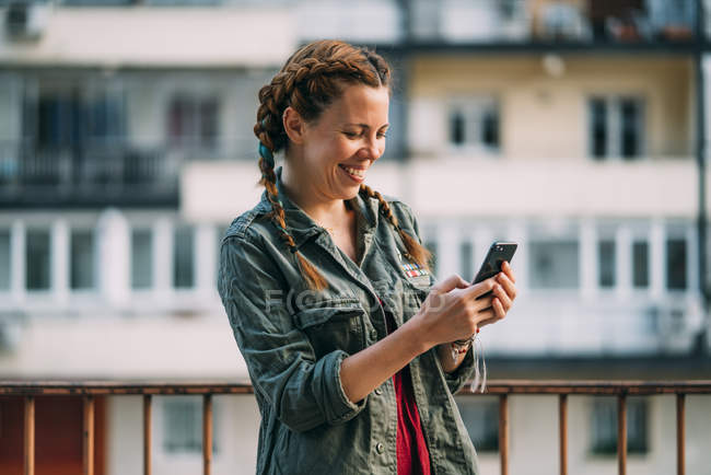 Смеющаяся рыжая девушка с косичками, использующая мобильный телефон против жилого дома — стоковое фото