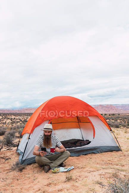 Bearded guy in casual outfit holding mug of hot beverage, salad and modern smartphone while sitting near tent and looking away in beautiful nature — Stock Photo