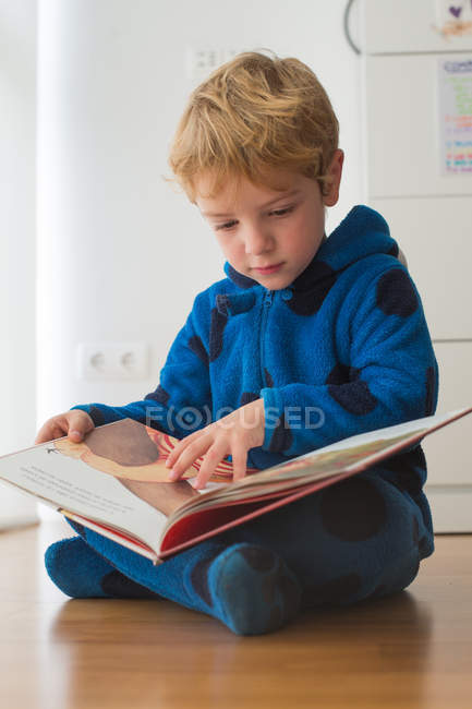 Focused blonde boy reading book on wooden floor — Stock Photo