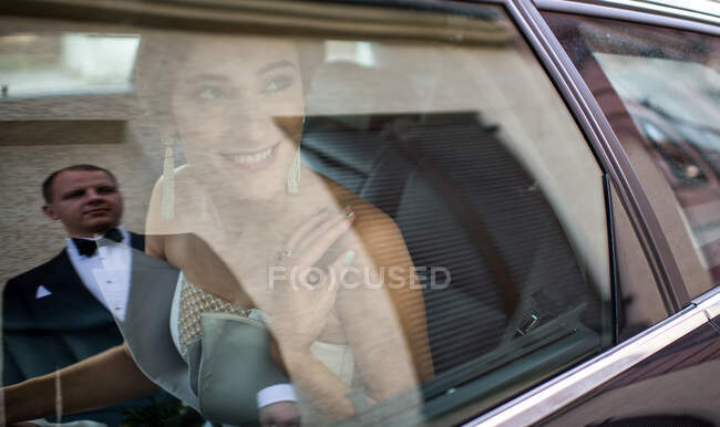 Belle jeune femme en robe de mariée souriant et regardant marié assis à l'intérieur de la voiture — Photo de stock