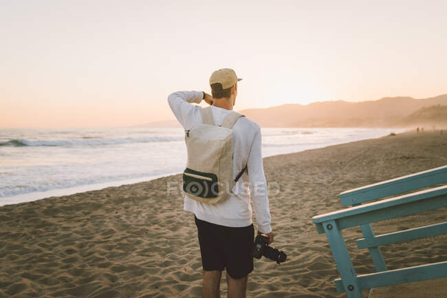 Вид на молодого парня с профессиональной фотокамерой, гуляющего по песчаному пляжу на закате в Санта-Монике, Калифорния — стоковое фото