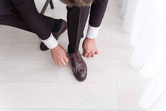 Jeune homme en costume noir et chemise blanche assis sur une chaise dans la chambre et portant des chaussures en cuir — Photo de stock