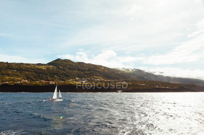 View of single ship with white sails flowing in blue water of sea against green mountains in sunlight — Stock Photo
