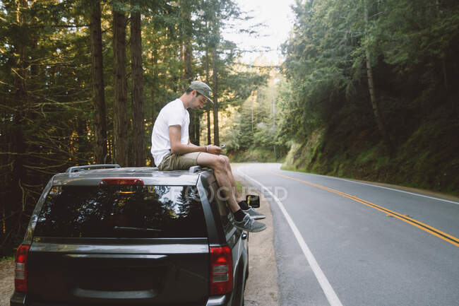 Side view of male in casual outfit browsing smartphone while sitting on car top on roadside in beautiful forest — Stock Photo