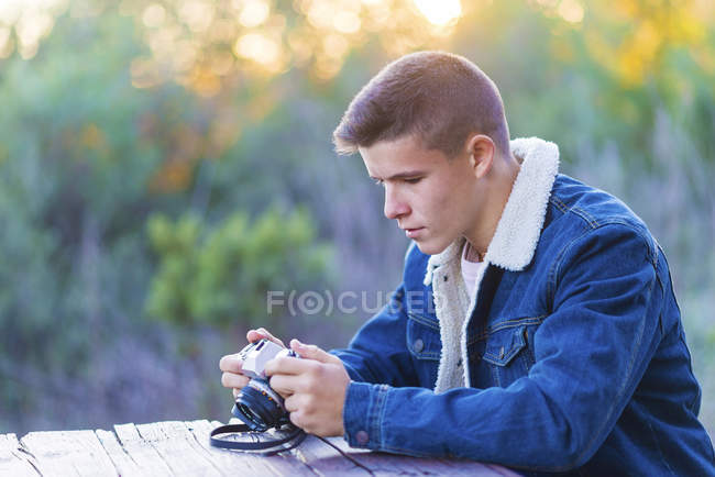 Молодой человек сидит за деревянным столом на улице с фотокамерой — стоковое фото