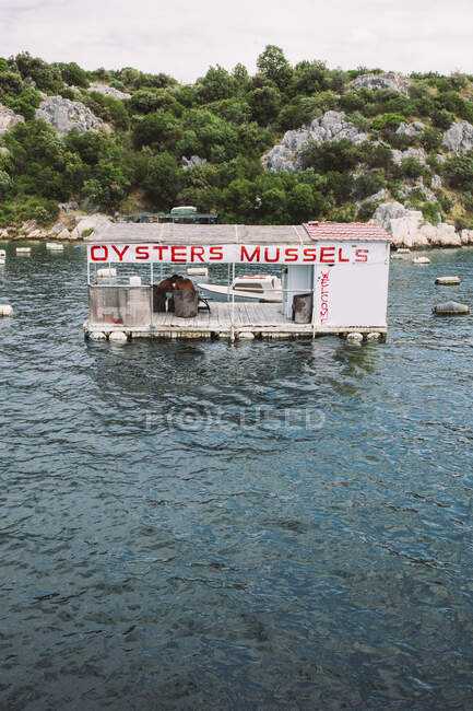 Blue rippling lake with little floating restaurant on background of rocks covered with green bushes — Stock Photo