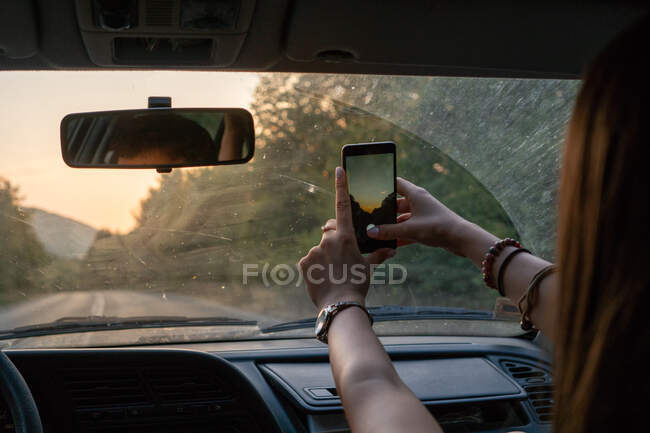 Неузнаваемая женщина, использующая смартфон для фотографирования, сидя на пассажирском сидении современного автомобиля во время поездки по Болгарии, на Балканах — стоковое фото