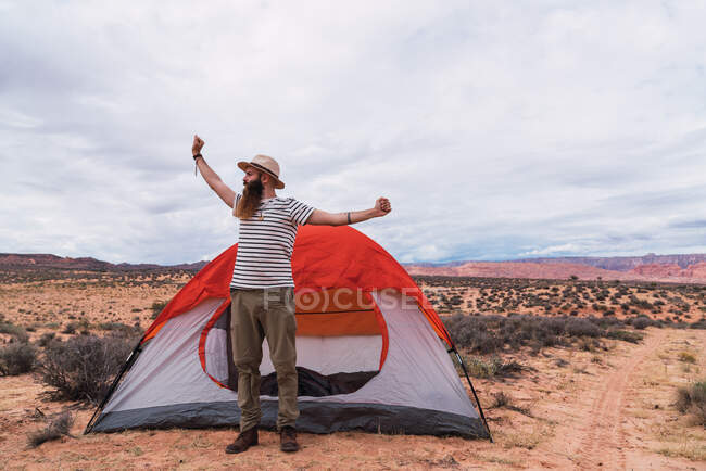 Нахабний бородатий самець розтягує руки і озирається, стоячи біля намету в похмурий день у дивовижній пустелі. — стокове фото