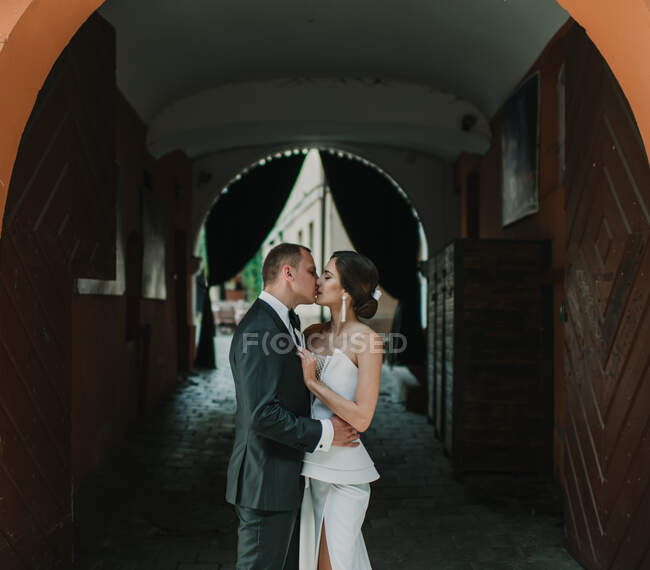 Vista lateral da jovem noiva e vassoura abraçando e beijando uns aos outros enquanto estão em pé no arco do edifício velho na rua da cidade — Fotografia de Stock