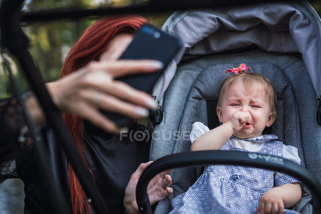 Madre mostrando smartphone a bebé disgustado en cochecito - foto de stock