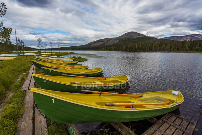 Barcos amarillos y verdes amarrados en la orilla del río ondulado en el fondo de las montañas y el cielo nublado - foto de stock