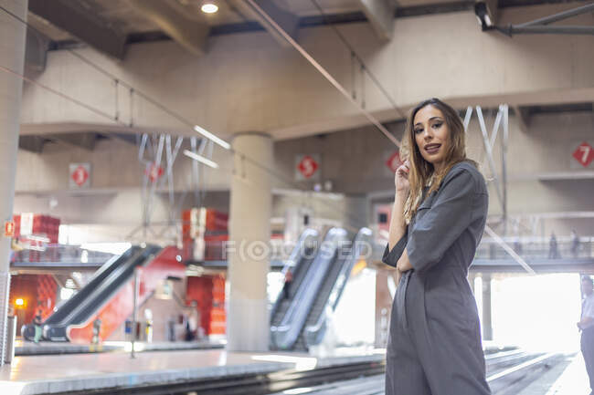 Вид позитивной дамы в элегантном наряде, улыбающейся и смотрящей в камеру, стоя на современном железнодорожном вокзале — стоковое фото