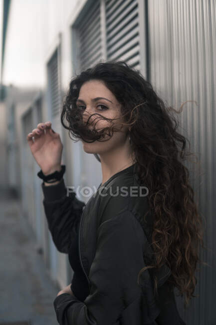 Side view of attractive young female with curly hair looking at camera while standing on city street on windy day — Stock Photo