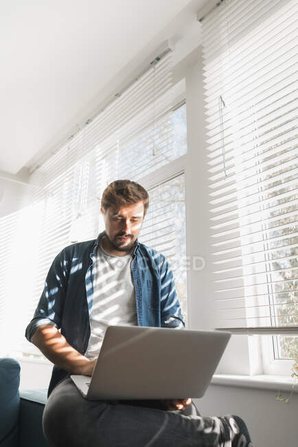 Бородатий хлопець переглядає сучасний ноутбук, сидячи на зручному дивані у стильній вітальні — стокове фото