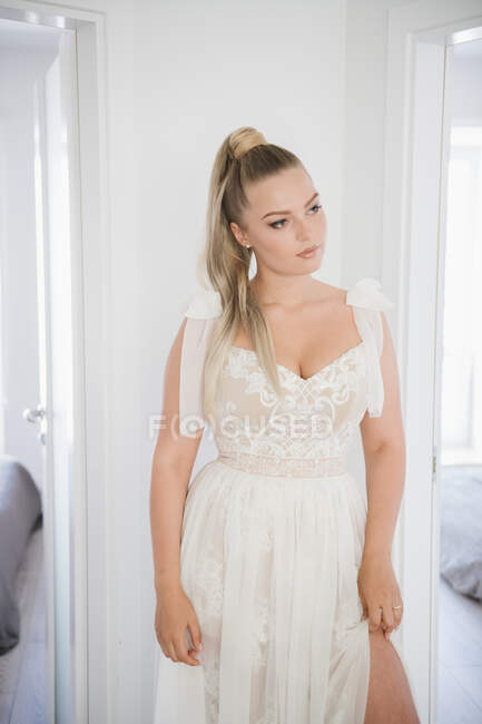 Jovem noiva bonita de pé e ajustando vestido branco enquanto olha para longe — Fotografia de Stock
