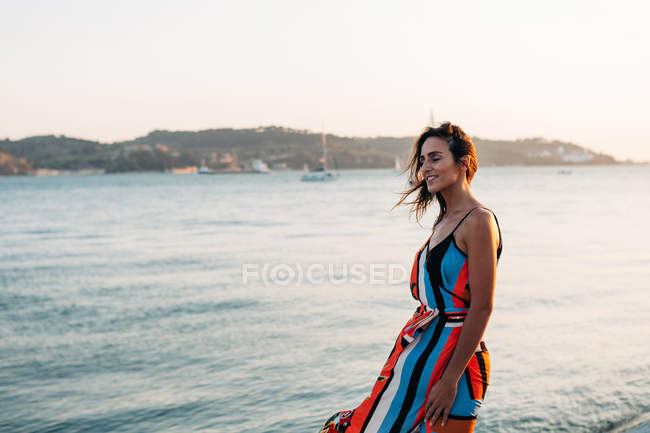 Zufriedene Frau in langem Kleid spaziert bei Sonnenuntergang über Kopfsteinpflaster-Promenade gegen das Meer — Stockfoto