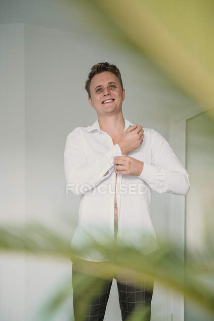 Handsome young man smiling and looking away while putting on white shirt before wedding ceremony — Stock Photo