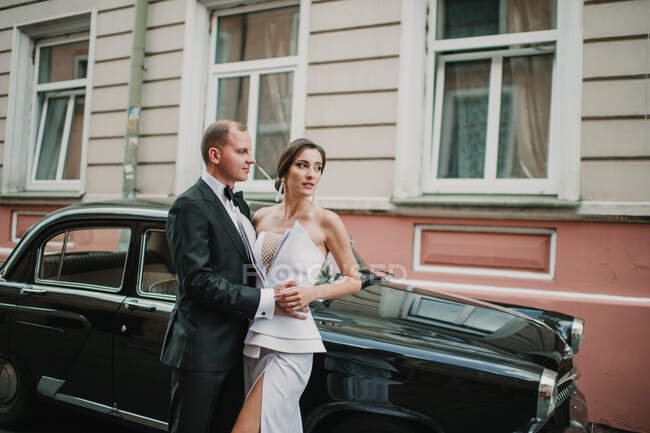 Vue latérale de beau jeune marié et jolie mariée étreinte tout en se tenant sur la rue de la ville près de véhicule vintage — Photo de stock