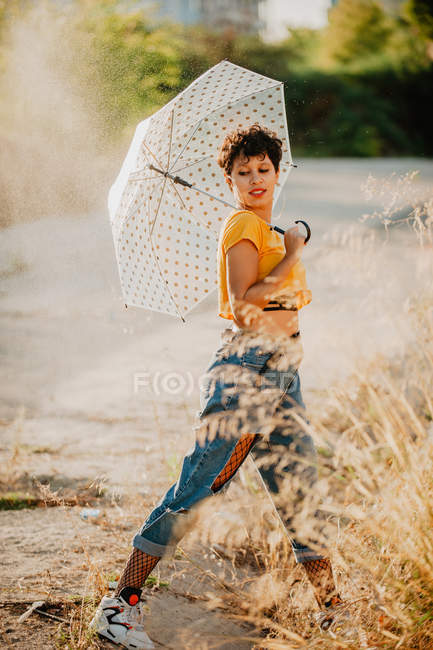 Mujer joven con paraguas en traje elegante sonriendo y mirando hacia otro lado mientras camina bajo gotas de agua de pulverización - foto de stock