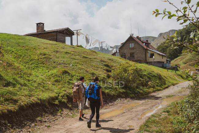 Visão traseira de duas pessoas com mochilas caminhando ao longo do caminho rural em direção a casas encantadoras no majestoso campo em dia nublado na Bulgária, Balcãs — Fotografia de Stock