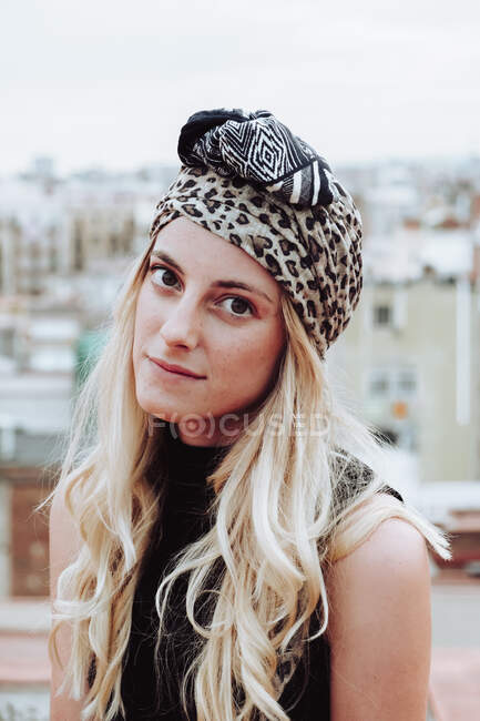 Jovem mulher atraente com cabelo loiro em pano de cabeça modelado sentado no telhado e olhando para a câmera no fundo da paisagem urbana — Fotografia de Stock