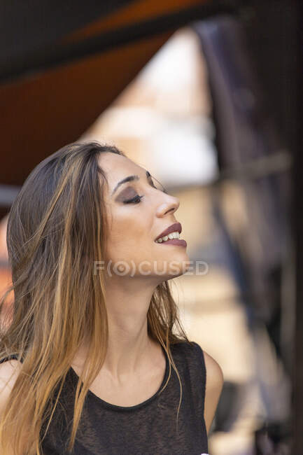 Vista lateral de la encantadora joven modelo sonriendo y mirando hacia otro lado mientras está de pie sobre el fondo borroso de la calle - foto de stock
