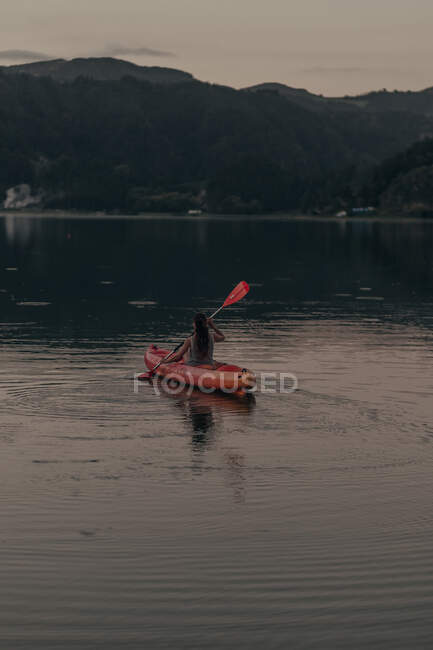 Вид ззаду на жінку, що пливе на човні в чистому нерухомому озері на тлі високих пагорбів і чистого неба — стокове фото