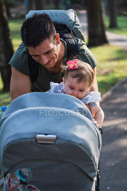 Handsome adult man smiling and putting sweet baby girl in carriage while standing on blurred background of park RELEASE — Stock Photo