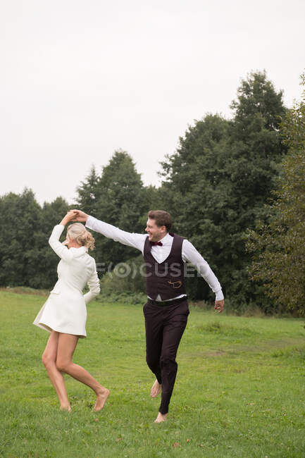 Trendy sposo adulto e sposa in abiti eleganti che si tengono per mano e saltano con eccitazione sul prato verde — Foto stock