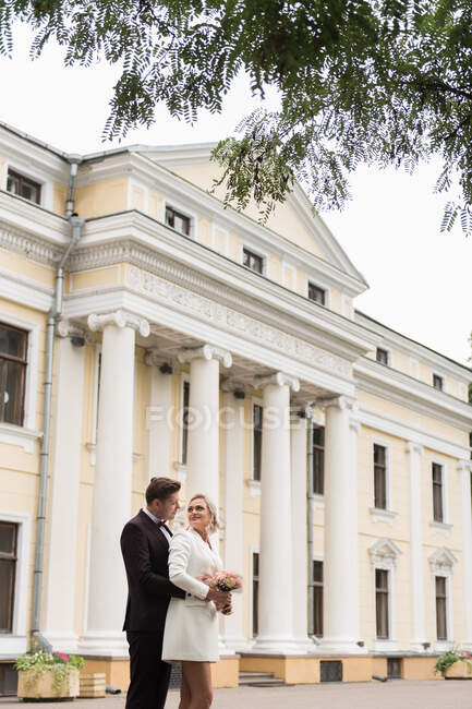 Снизу красивая женщина и мужчина в элегантных свадебных костюмах и улыбаясь друг другу на фоне старого здания — стоковое фото
