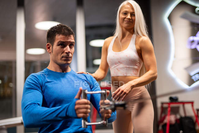 Чоловік робить вправи в спортзалі з жінкою, що стоїть і посміхається — стокове фото