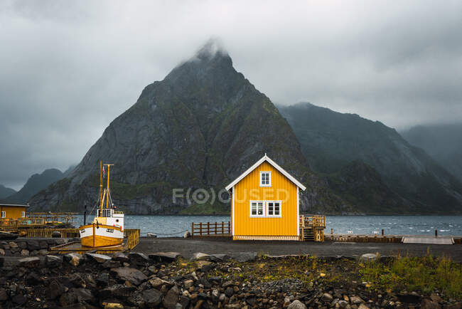 Landscape of little yellow house standing on coast of blue river on background of high mountains covered with fog — Stock Photo
