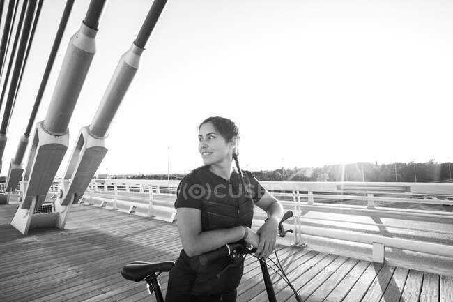 Felice donna sportiva con bicicletta moderna in piedi sulla costruzione pavimentata di passerella alla luce del sole — Foto stock