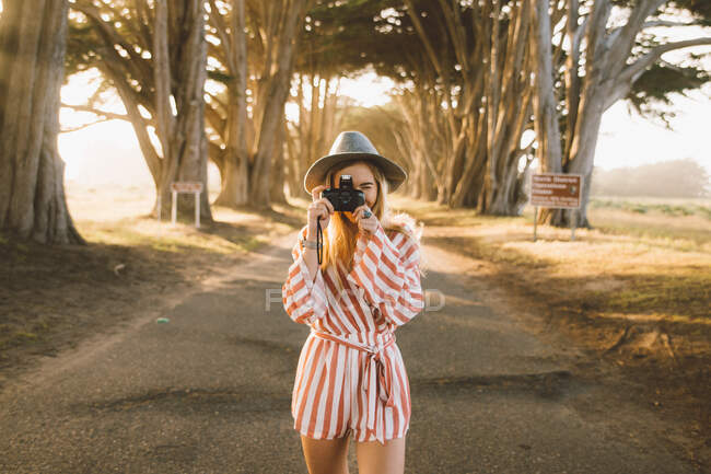 Молода жінка в стильному вбранні, використовуючи фотоапарат, щоб сфотографуватися, стоячи на дорозі в дивовижному дерев'яному тунелі в сонячний день у чудовій природі — стокове фото