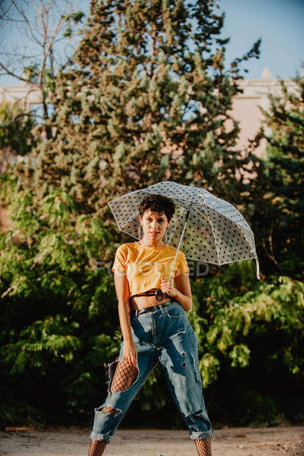 Attraktive junge Frau in stylischem, lässigem Outfit, die in die Kamera schaut, während sie einen Regenschirm hält und in der Nähe von Büschen auf der Straße steht — Stockfoto