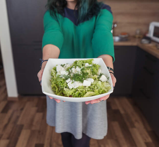 Кукурудза жінка в повсякденному одязі демонструє миску зі свіжим салатом до камери, стоячи на стильній кухні вдома — стокове фото