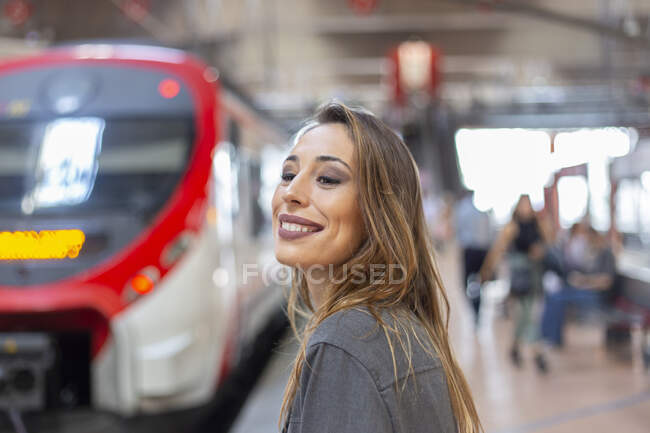 Vista lateral da linda fêmea sorrindo e olhando para longe enquanto estava no fundo da moderna estação ferroviária — Fotografia de Stock