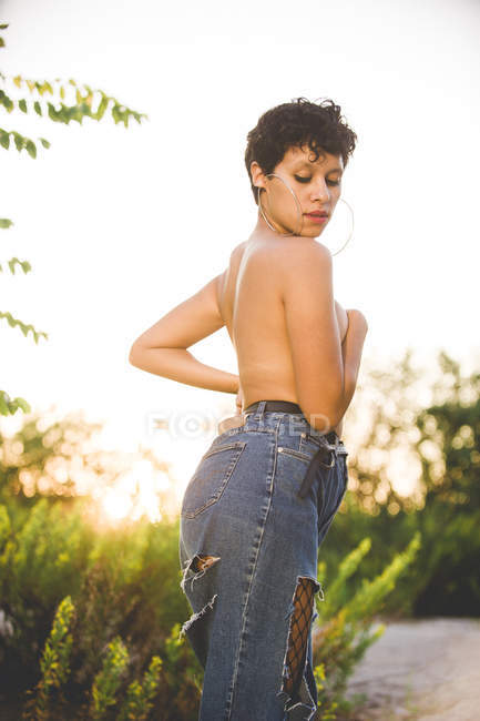 Уверенная провокационная женщина в джинсе, стоящая топлесс, прикрывающая грудь в природе — стоковое фото