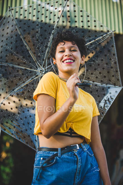Портрет молодой женщины с прозрачным зонтиком, улыбающейся и смотрящей в камеру, стоя на улице — стоковое фото