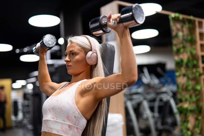 Young blond woman in sport top with headset doing exercise with iron dumbbells on gym equipment — Stock Photo