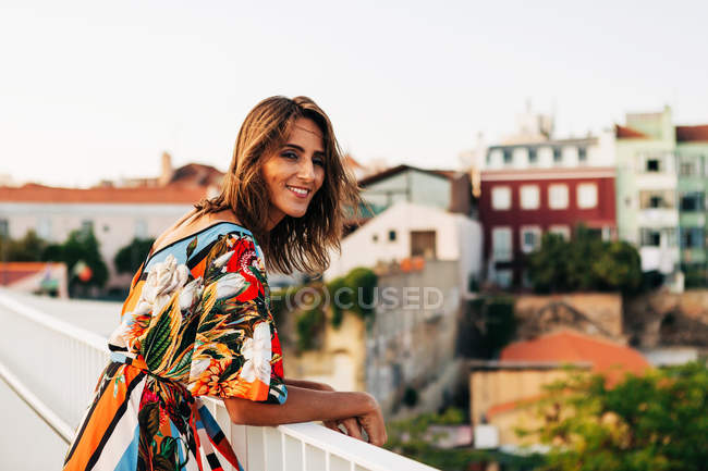 Happy brunette woman in colorful dress standing on bridge and looking at camera on city background — Stock Photo