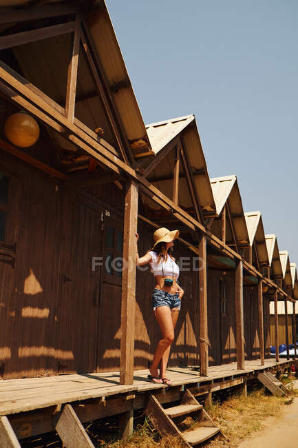Неузнаваемая загорелая женщина с каштановыми волосами и соломенной шляпой в джинсовых шортах и белом топе, стоящая и опирающаяся на деревянное здание на пляже — стоковое фото