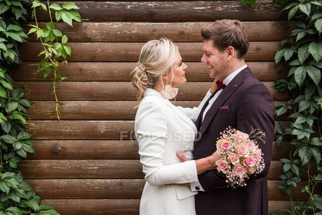 Elegant adult bride and groom embracing while standing in suits against wooden wall and smiling at camera — Stock Photo