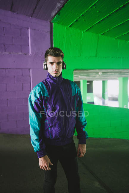 Jovem homem bonito em casaco esporte com fone de ouvido em pé no fundo da parede colorida e olhando para a câmera — Fotografia de Stock