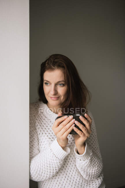 Hermosa hembra adulta en suéter de punto sonriendo y mirando hacia otro lado mientras sostiene la taza de bebida caliente fresca y apoyada en la pared - foto de stock