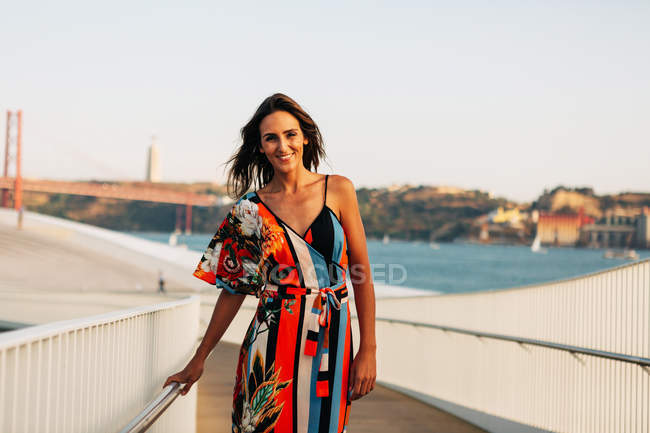 Elegante lächelnde Frau im Kleid, die im Sonnenlicht auf einer Brücke spaziert — Stockfoto