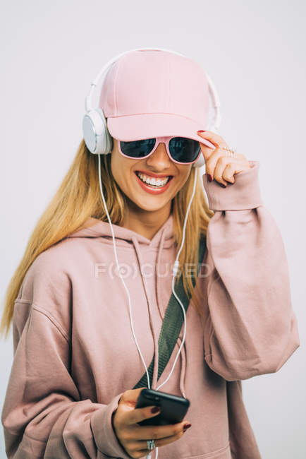 Mujer elegante con capucha rosa y gorra escuchando música con auriculares - foto de stock