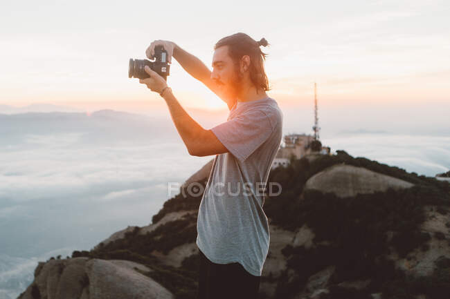 Side view of handsome bearded guy using professional photo camera to take photos on amazing nature while standing on mountain during sunrise in Barcelona, Spain — Stock Photo