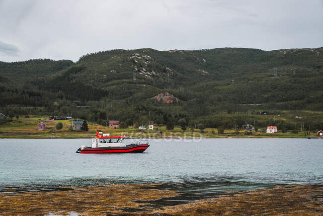 Маленький червоний моторний човен, що плаває на спокійній блакитній річці на тлі мальовничих зелених пагорбів і чистого неба — стокове фото